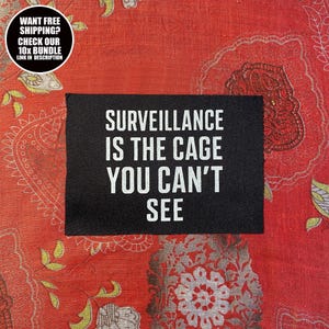 May include: A black patch with white text reading "SURVEILLANCE IS THE CAGE YOU CAN'T SEE" on a red patterned background. A circular graphic in the top left corner says "WANT FREE SHIPPING? CHECK OUR 10X BUNDLE LINK IN DESCRIPTION."