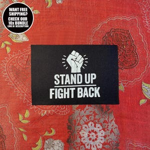 Puede incluir: Un parche rectangular negro con las palabras "STAND UP FIGHT BACK" en blanco, debajo de un símbolo de puño blanco. El parche está sobre un fondo rojo estampado. Un gráfico circular en la esquina superior izquierda dice "WANT FREE SHIPPING? CHECK OUR 10X BUNDLE LINK IN DESCRIPTION."