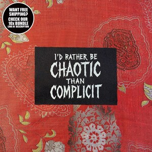 Op de afbeelding: Een zwart rechthoekig embleem met de witte tekst "I'D RATHER BE CHAOTIC THAN COMPLICIT". Het embleem is geplaatst op een rode stof met een patroon. Een cirkelvormige afbeelding linksboven luidt "WANT FREE SHIPPING? CHECK OUR 10X BUNDLE LINK IN DESCRIPTION."