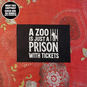 Könnte beinhalten: Schwarzer rechteckiger Aufnäher mit weißem Text: „A ZOO IS JUST A PRISON WITH TICKETS“. Das Design zeigt eine Silhouette einer Person hinter Gittern. Auf rotem Stoff mit Muster.