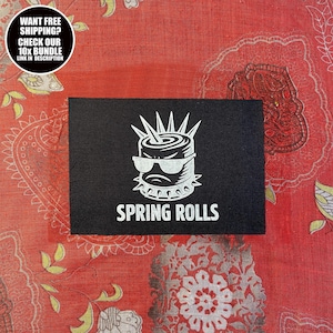 May include: A black rectangular patch featuring a white graphic of a spring roll with sunglasses and a spiky mohawk. The text "SPRING ROLLS" is below the graphic. The background is a red patterned fabric.