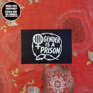 Op de afbeelding: Een zwarte patch met de witte tekst "GENDER IS A PRISON" in een cirkel van prikkeldraad. De patch heeft ook een vrouwelijk symbool en verticale lijnen. De achtergrond is een rode stof met een patroon.
