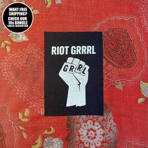 May include: Black rectangular patch with the words "RIOT GRRRL" in white block letters. Below the text is a white fist with the letters "GRRL" on the knuckles. The patch is on a red patterned background.