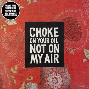 May include: A black square patch with white text that reads "CHOKE ON YOUR OIL NOT ON MY AIR." The patch is set against a red patterned background. A circular text bubble in the upper left corner says "WANT FREE SHIPPING? CHECK OUR 10X BUNDLE LINK IN DESCRIPTION."