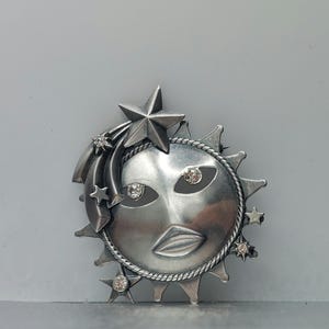 May include: A silver sun face brooch with a shooting star and rhinestones. The sun face has a stylized face with a mouth and eyes.