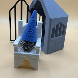 May include: A blue and white 3D printed dice tower with a blue and white dice and a blue cone with the word "DUNCE" printed on it. The tower has a gray roof with the word "LAME" printed on it.