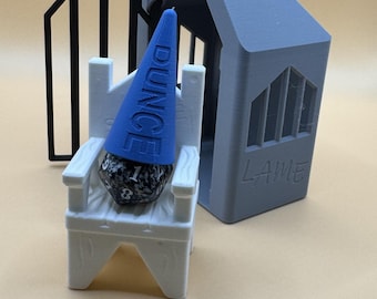 May include: A blue and white 3D printed dice tower with a blue and white dice and a blue cone with the word "DUNCE" printed on it. The tower has a gray roof with the word "LAME" printed on it.