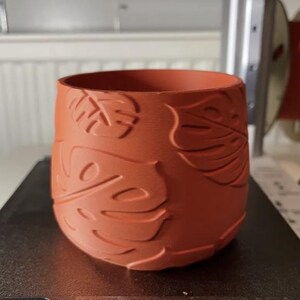 May include: A terracotta-colored cylindrical planter with a textured surface featuring embossed monstera leaves. The planter is designed with a wide opening and a rounded base. The product is suitable for indoor or outdoor use.