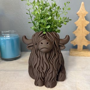 May include: A brown Highland cow plant pot with a shaggy coat, filled with green foliage. The pot features curved horns and a detailed face. A blue candle and wooden tree are in the background.