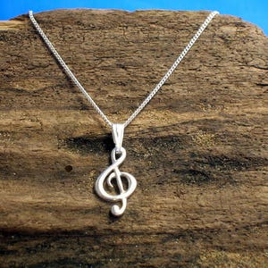 May include: A silver chain necklace with a treble clef pendant. The necklace is displayed on a piece of driftwood.