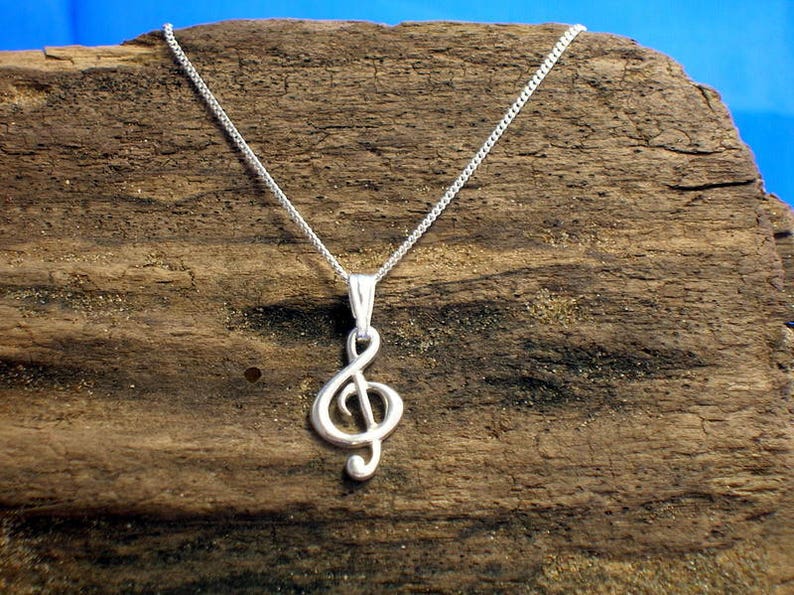 May include: A silver chain necklace with a treble clef pendant. The necklace is displayed on a piece of driftwood.