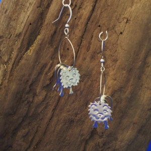 May include: A pair of silver earrings with a whimsical design. Each earring features a small, round, silver charm with a face and a textured surface. The charms are attached to a silver hoop.