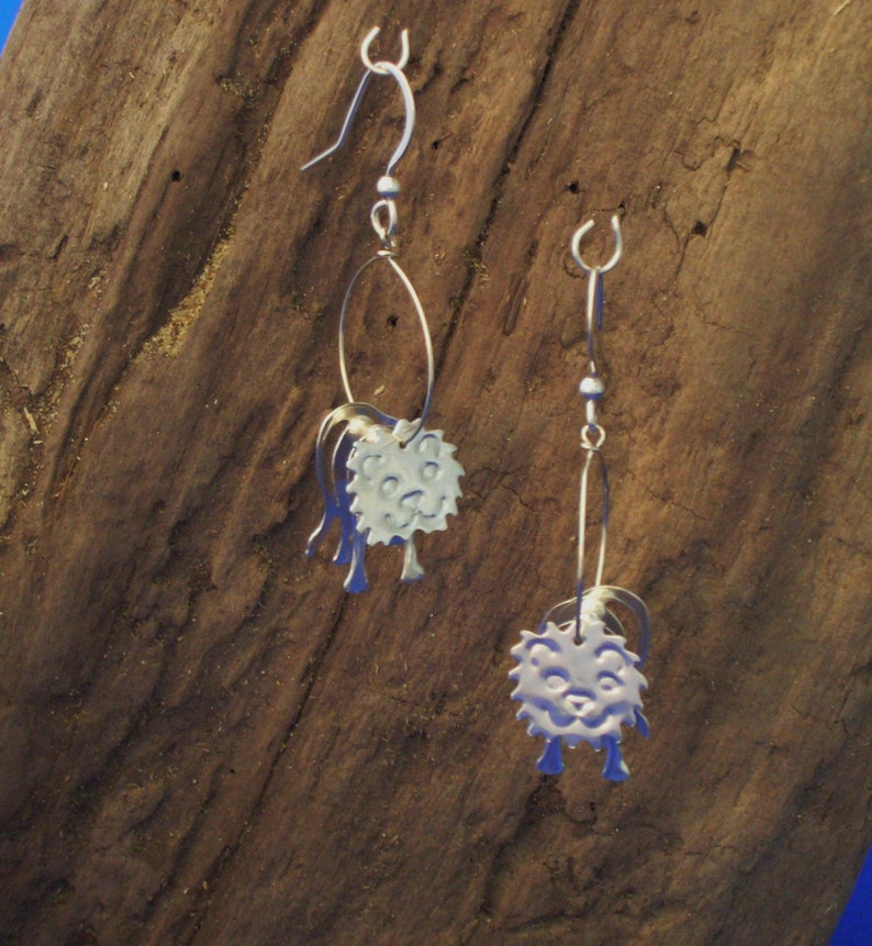 May include: A pair of silver earrings with a whimsical design. Each earring features a small, round, silver charm with a face and a textured surface. The charms are attached to a silver hoop.