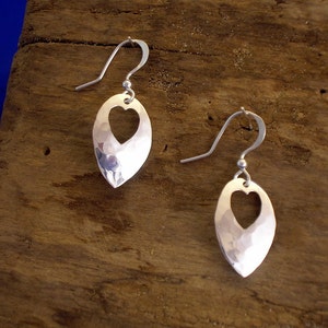 May include: A pair of silver drop earrings. Each earring has a hammered, oval form with a heart-shaped cut-out. The earrings have a simple hook fastening.