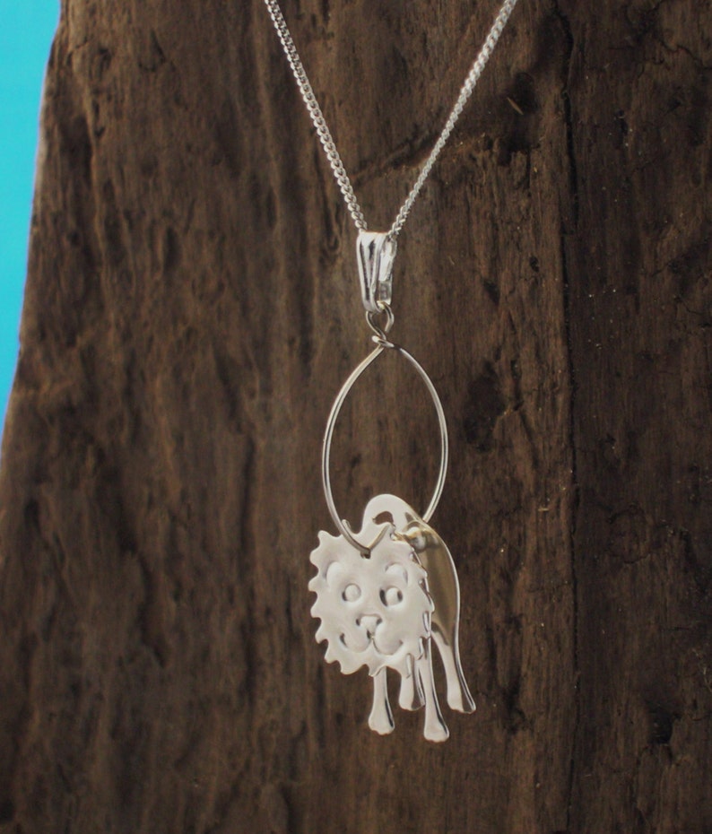 May include: A silver necklace with a charm of a cartoon lion with a happy expression. The charm is attached to a silver chain with a silver loop.