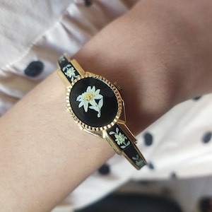 May include: A gold-toned bracelet with a black oval centerpiece featuring a white flower design. The bracelet has black and gold accents with floral patterns. The watch is worn on a wrist, showcasing the intricate details of the jewelry.