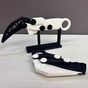 May include: A 3D-printed, black and white claw-shaped object, with a curved blade and a ring handle. The claw is displayed on a black stand, with a second claw lying flat in front. The design features geometric shapes and a mechanical aesthetic.