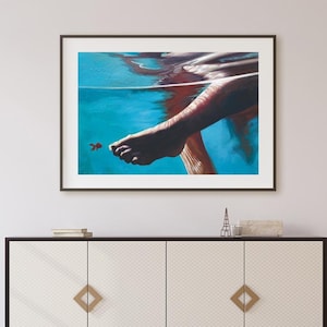 Puede incluir: Una impresión artística enmarcada muestra una pierna y un pie bajo el agua, con un pequeño pez naranja. El agua es de un azul vibrante. La obra de arte cuelga sobre un mueble blanco y negro con detalles dorados.