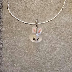 May include: A silver necklace featuring a pendant shaped like a bunny. The bunny pendant is white with pink accents on the ears and a smiling face. The necklace is displayed on a textured, light gray surface.
