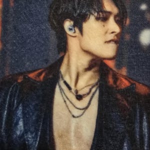 May include: A portrait of a person with dark hair and a black jacket. The person is wearing layered necklaces. The background is blurred with warm tones.