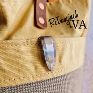 Puede incluir: Primer plano de una bolsa de lona con un cierre metálico. La bolsa es una combinación de tela beige y gris, con una correa de cuero marrón y el texto "ReImagined in VA". El cierre es plateado y curvado.