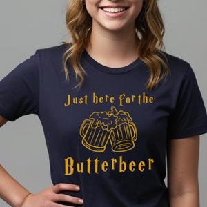 May include: Navy blue t-shirt with the text "Just here for the Butterbeer" in gold. The design features two mugs of frothy beer. A casual, comfortable tee for fans of a popular fantasy series.