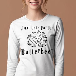 以下が含まれることがあります： 「Just here for the Butterbeer」の黒い文字が入った白い長袖シャツ。泡立ったビールが入ったマグカップが2つ描かれています。柔らかい素材のシャツです。