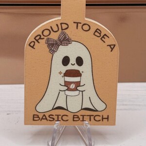 May include: A tan-colored bookmark featuring a cartoon ghost with a bow holding a coffee cup. The text "PROUD TO BE A BASIC BITCH" is printed on the bookmark. The bookmark is displayed on a clear plastic stand.