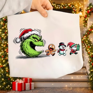 Könnte beinhalten: Weiße Weihnachtstasche mit einem grünen Grinch mit Weihnachtsmütze, der einen Lebkuchenmann, einen Schneemann und einen Elfen jagt. Der Grinch hat einen wütenden Gesichtsausdruck. Die anderen Charaktere rennen weg. Die Tasche wird von einer Person gehalten.