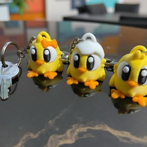 May include: Three yellow chick keychain charms with black eyes and orange feet. The charms are attached to silver key rings.