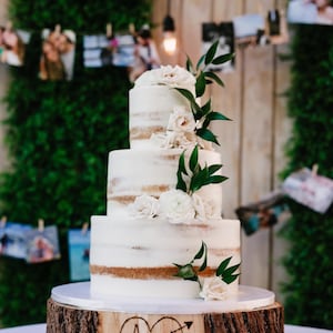 K&ouml;nnte beinhalten: Eine wei&szlig;e Hochzeitstorte mit vier Etagen, verziert mit wei&szlig;en Blumen und Gr&uuml;n. Die Torte steht auf einem Holzkuchenst&auml;nder mit den Initialen "A + B", die in das Holz geschnitzt sind.
