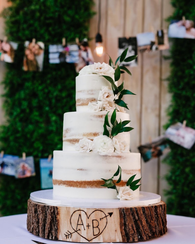 K&ouml;nnte beinhalten: Eine wei&szlig;e Hochzeitstorte mit vier Etagen, verziert mit wei&szlig;en Blumen und Gr&uuml;n. Die Torte steht auf einem Holzkuchenst&auml;nder mit den Initialen "A + B", die in das Holz geschnitzt sind.