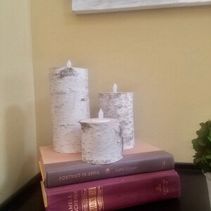 May include: Three white birch bark pillar candles with flickering LED lights, arranged on a stack of books. The books have titles "Portrait in Sepia" and "James A. Michener" visible.