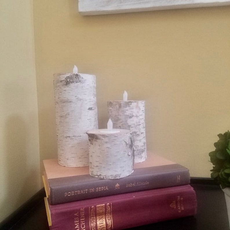 May include: Three white birch bark pillar candles with flickering LED lights, arranged on a stack of books. The books have titles "Portrait in Sepia" and "James A. Michener" visible.