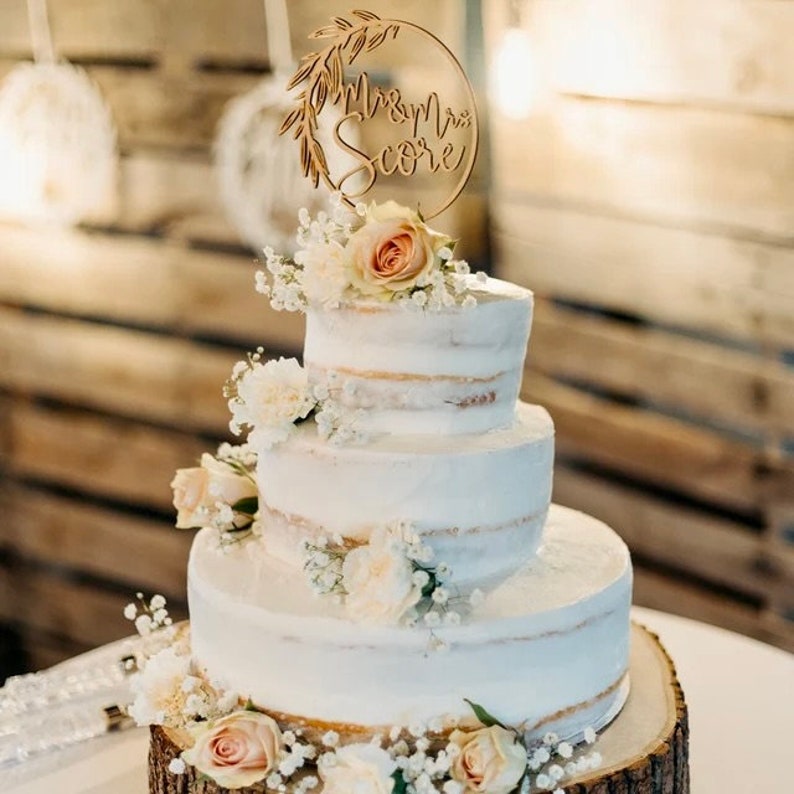 May include: A three-tiered wedding cake decorated with white frosting and fresh flowers. The cake is topped with a wooden cake topper that reads "Mr & Mrs Score".