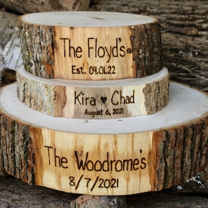 K&ouml;nnte beinhalten: Drei Holzkuchenst&auml;nder mit Rindenkanten. Der obere St&auml;nder ist mit "The Floyds" und "Est. 04.01.22" graviert. Der mittlere St&auml;nder ist mit "Kira &hearts; Chad" und "August 6, 2021" graviert. Der untere St&auml;nder ist mit "The Woodromes" und "8/7/2021" graviert.