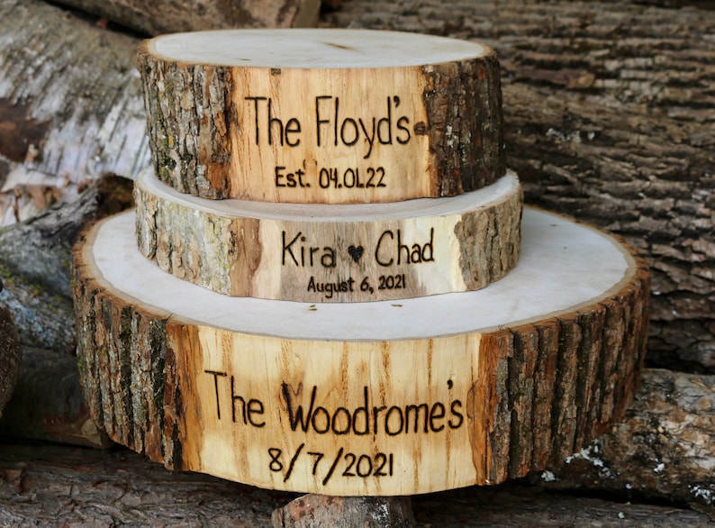 K&ouml;nnte beinhalten: Drei Holzkuchenst&auml;nder mit Rindenkanten. Der obere St&auml;nder ist mit "The Floyds" und "Est. 04.01.22" graviert. Der mittlere St&auml;nder ist mit "Kira &hearts; Chad" und "August 6, 2021" graviert. Der untere St&auml;nder ist mit "The Woodromes" und "8/7/2021" graviert.