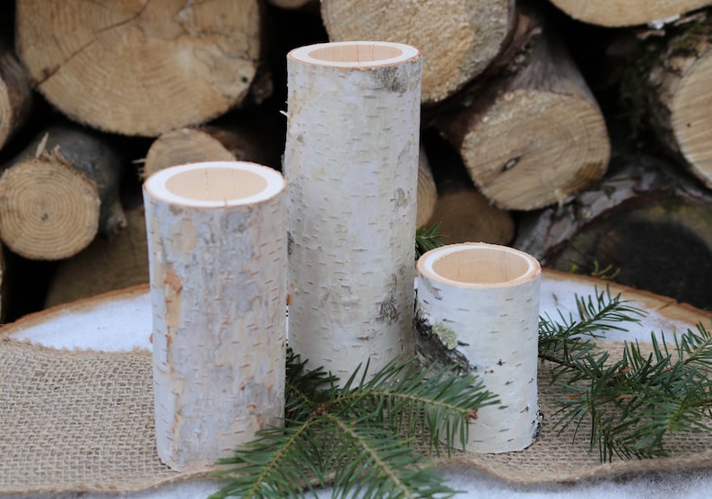 May include: Three white birch bark candle holders of varying sizes. The candle holders are sitting on a burlap surface with a pine bough in front of them.