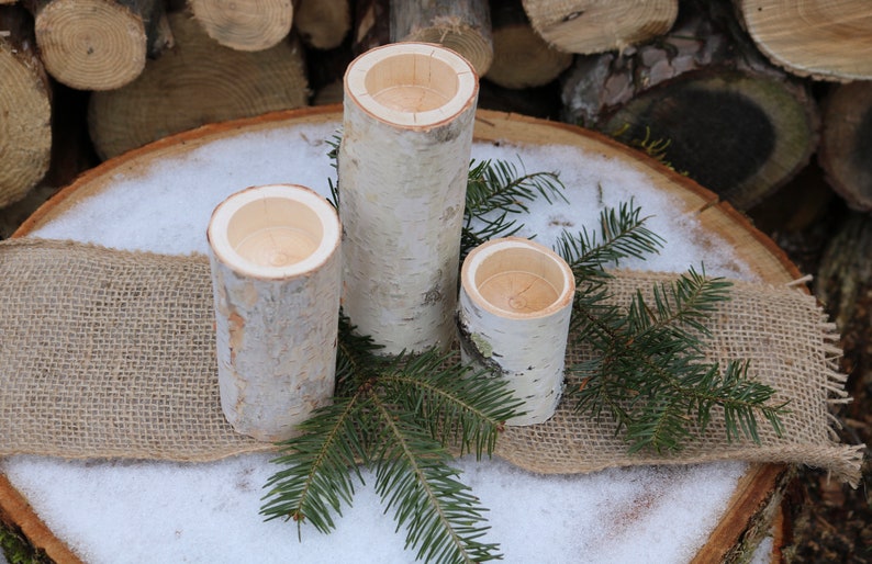 May include: Three white birch bark candle holders of varying sizes, arranged on a wooden slice with burlap and evergreen sprigs.