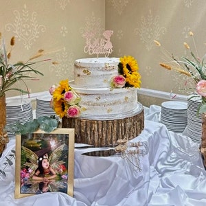 May include: A two-tiered white cake with gold accents and a wooden base. The cake is decorated with pink and yellow flowers and a topper that says "Mi Bautizo". The cake is on a white tablecloth with two pairs of cowboy boots and a framed photo of a young girl.