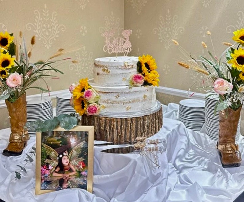 May include: A two-tiered white cake with gold accents and a wooden base. The cake is decorated with pink and yellow flowers and a topper that says "Mi Bautizo". The cake is on a white tablecloth with two pairs of cowboy boots and a framed photo of a young girl.