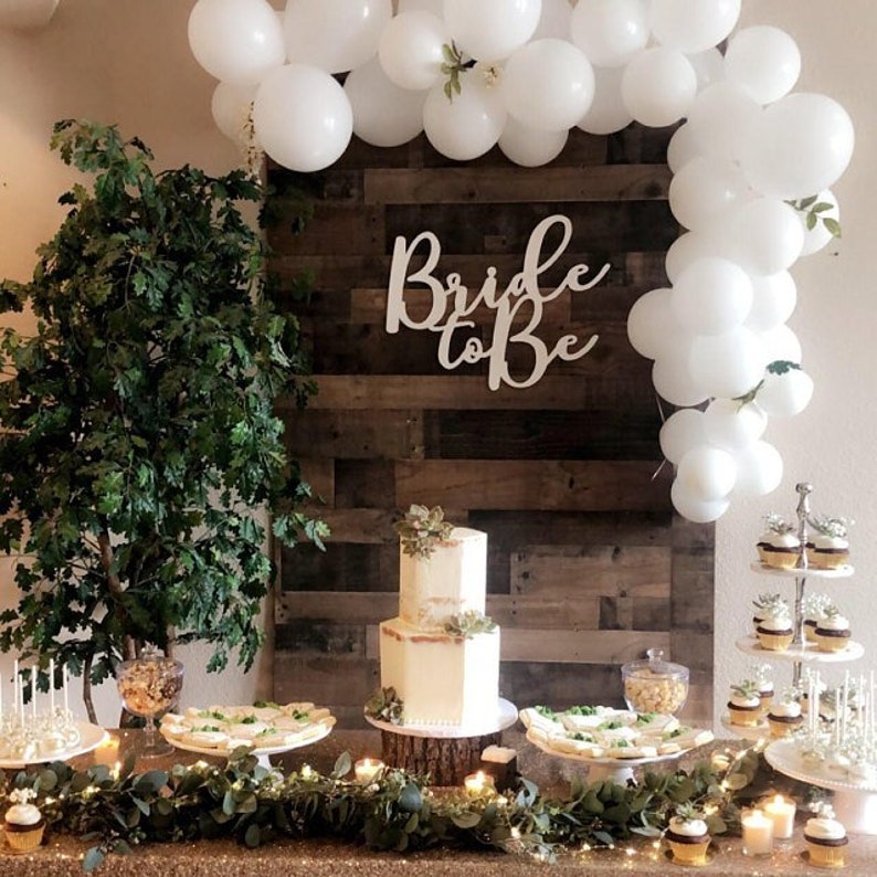 May include: A bridal shower dessert table with a white cake decorated with greenery, cupcakes, and other treats. The backdrop is a rustic wood wall with a white sign that says "Bride to Be" and a white balloon garland.