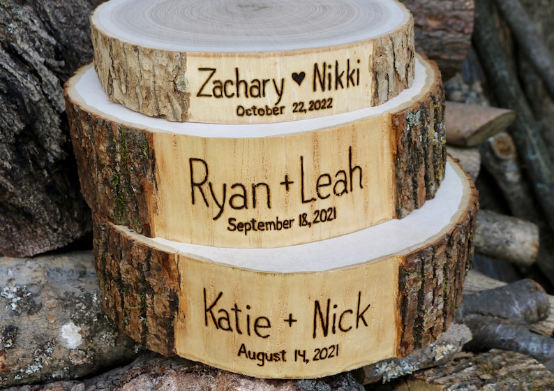 K&ouml;nnte beinhalten: Drei Holzkuchenst&auml;nder mit Rindenkanten, &uuml;bereinander gestapelt. Der obere St&auml;nder ist mit "Zachary Nikki October 22, 2022" graviert. Der mittlere St&auml;nder ist mit "Ryan + Leah September 18, 2021" graviert. Der untere St&auml;nder ist mit "Katie + Nick August 14, 2021" graviert.