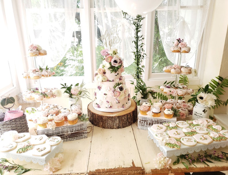 May include: A pink and white tiered cake with floral decorations sits on a wooden slice. The cake is surrounded by cupcakes, donuts, and cookies, all decorated with pink and white frosting and flowers. The table is covered in a white tablecloth and there are green plants in the background.