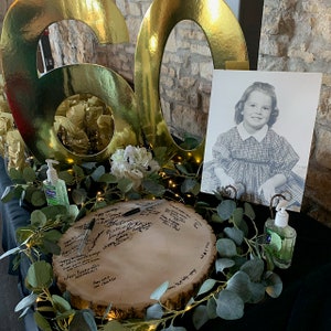 May include: A gold 60th birthday sign with a black and white photo of a young girl in a plaid dress. The photo is on a white card and is propped up on a table. There is a wooden slice with signatures on it, surrounded by greenery and string lights. There are also two clear bottles with pumps, filled with hand sanitizer.