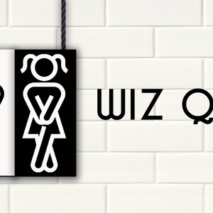 Puede incluir: Un letrero en blanco y negro con un símbolo de baño para hombres y mujeres colgado en una pared de ladrillo blanco. El letrero dice "WIZ QUIZ".