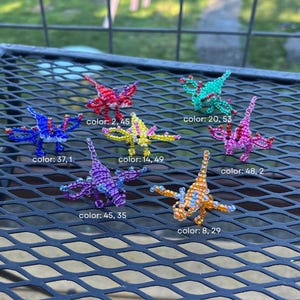 May include: A collection of seven beaded dragonfly ornaments in various colors, including red, blue, yellow, green, pink, purple, and orange. Each dragonfly has a unique color combination and is displayed on a black metal surface.
