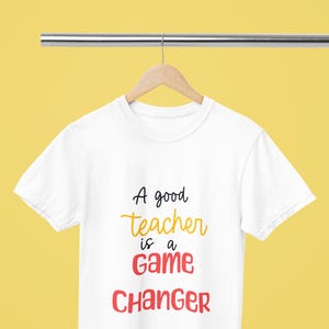 May include: White t-shirt with the text "A good teacher is a game changer" in black, yellow, and red lettering.