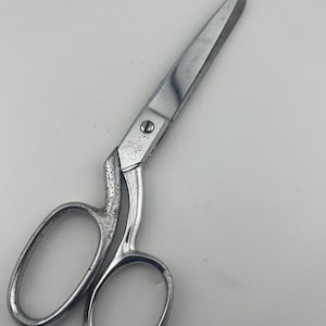 May include: Vintage Singer scissors with a silver finish. The scissors have large finger holes and a curved handle. The blades are pointed and show the Singer brand name. The scissors are likely used for sewing or fabric cutting.