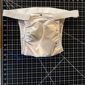 May include: A beige cloth diaper with a white waistband and adjustable straps. The diaper has a snap closure and is laid out on a black cutting mat with a grid pattern. A blue elastic band is also visible.